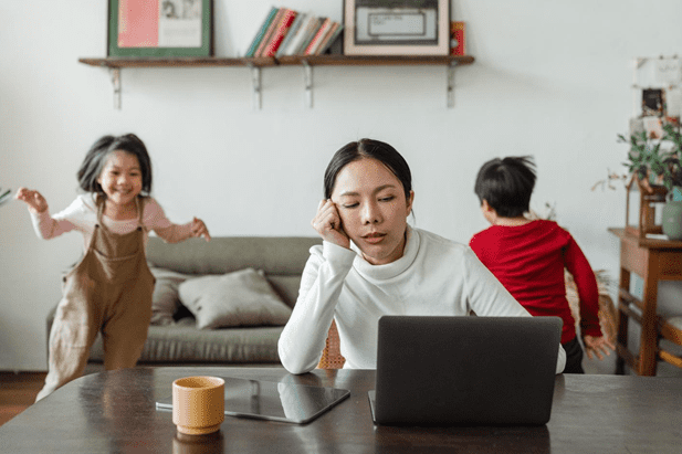 Stressed mom working at home - Woman sitting in front of a laptop looking sad and tired while her two kids and running and jumping around her