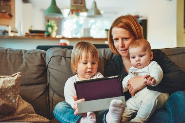 Photo of a woman holding her two kids and looking up something on a tablet