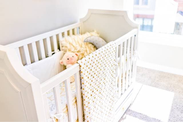 A close-up of a white crib with a pillow and stuffed animals inside and a polka dot blanket hanging on the side.